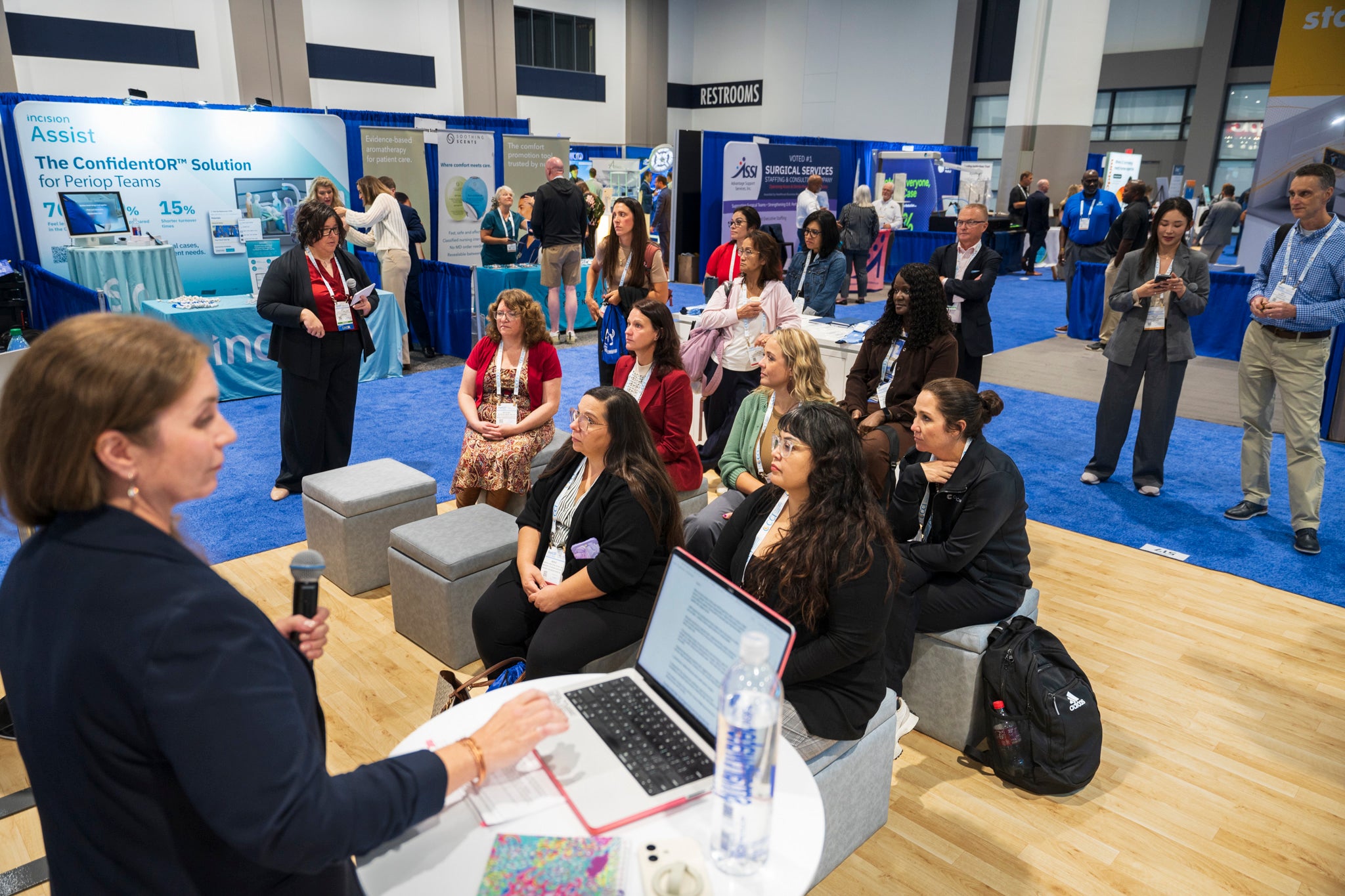Attendees seated on cubes listening to a presenter on the Hall floor, surrounded by exhibitor booths